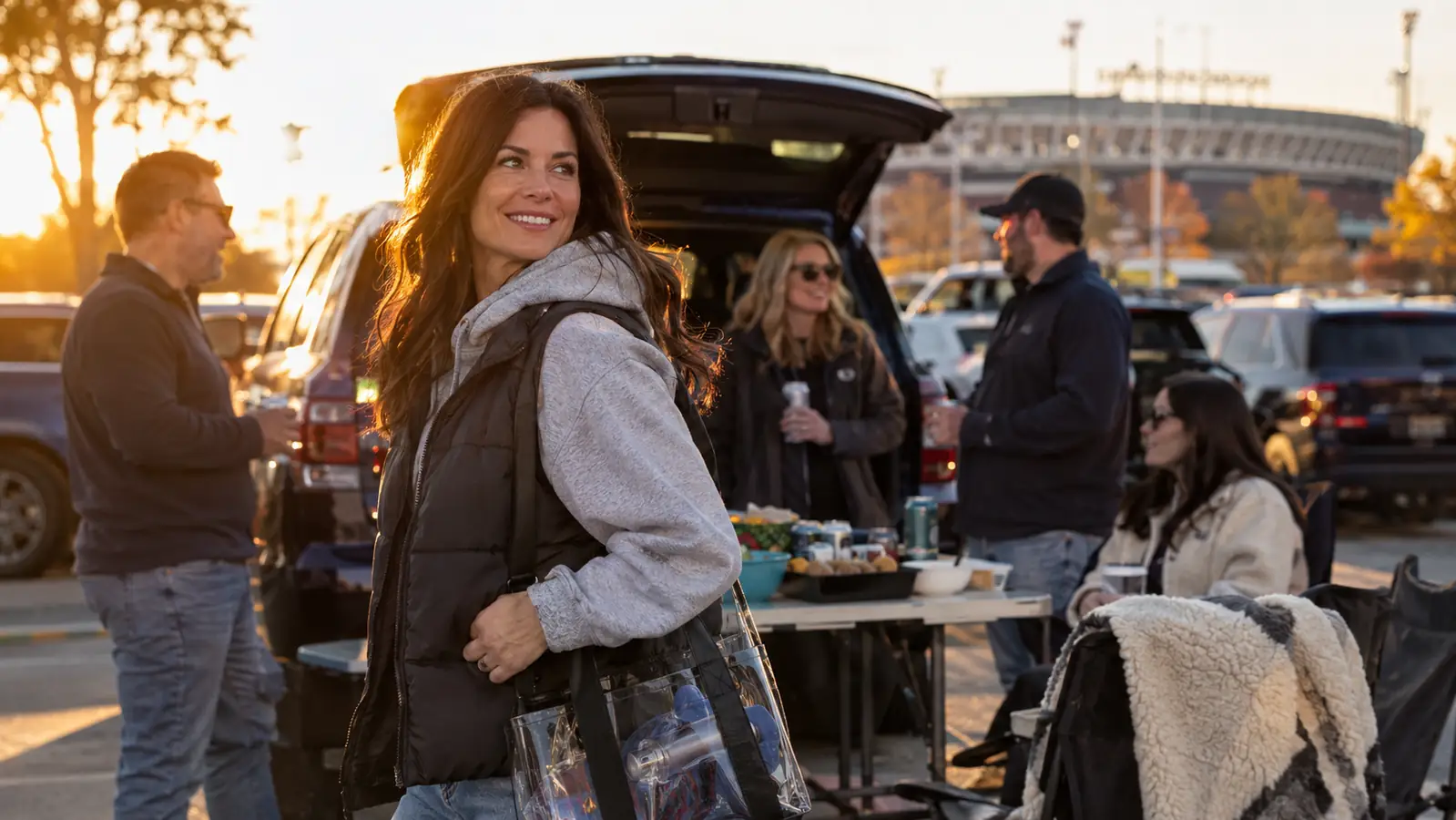 A smiling woman in a cozy hoodie and heated vest holding a clear stadium tote at an autumn tailgate beside an open SUV