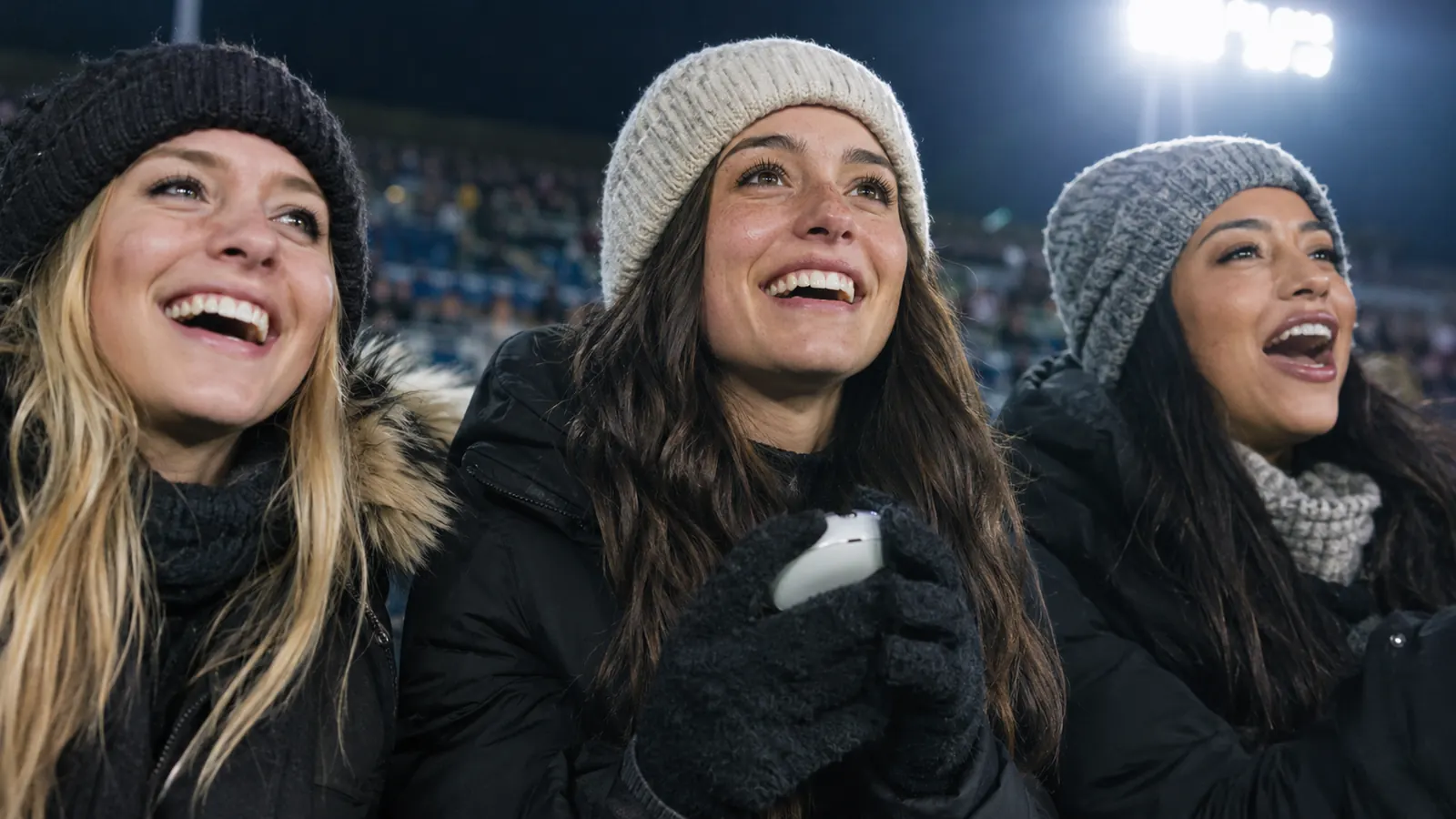 A woman on a stadium seat between two friends, wearing gloves while holding a small device, everyone cheering, cold evening lights