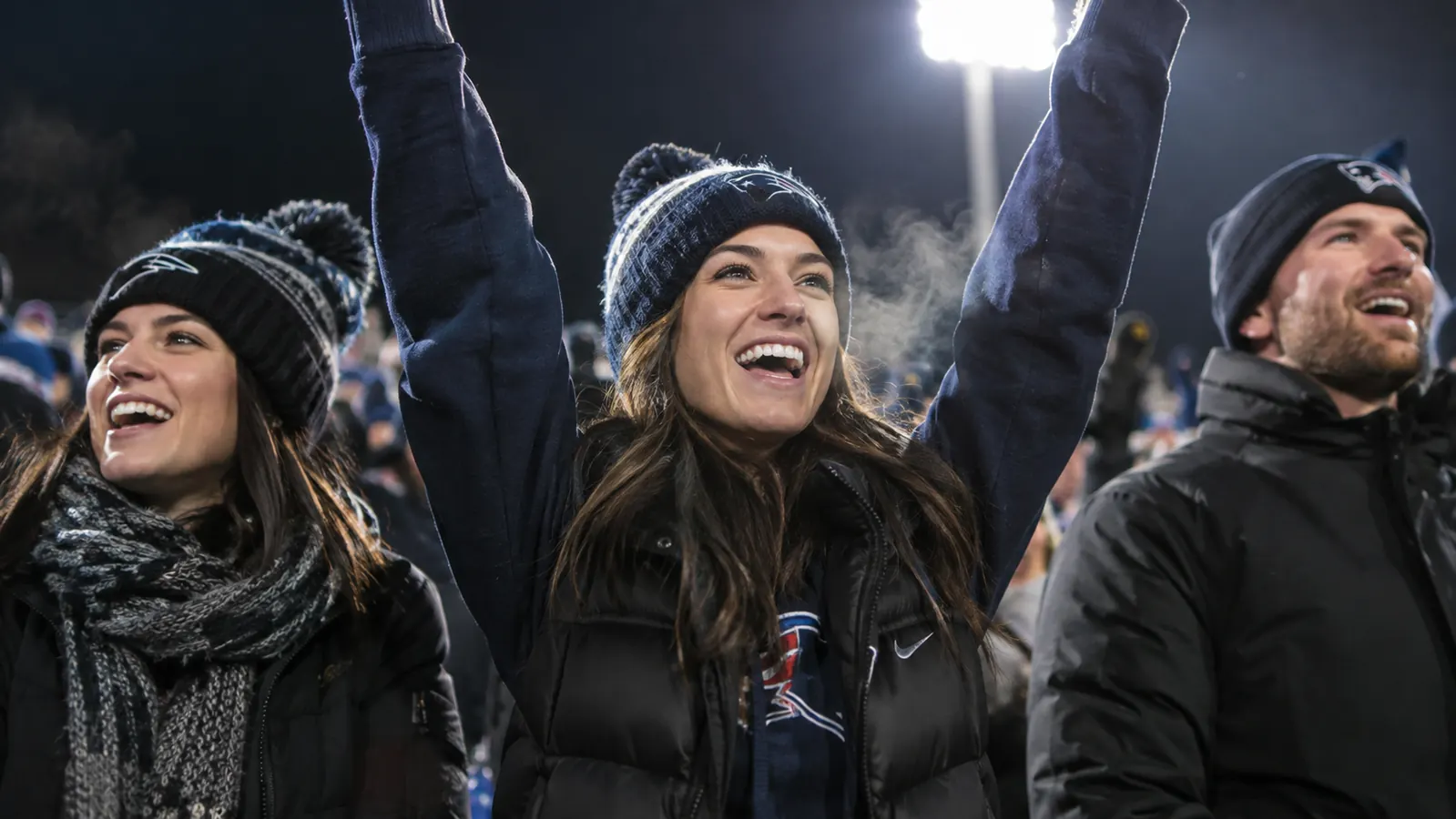 A woman in a team hoodie and heated vest raising both arms and cheering in a cold stadium