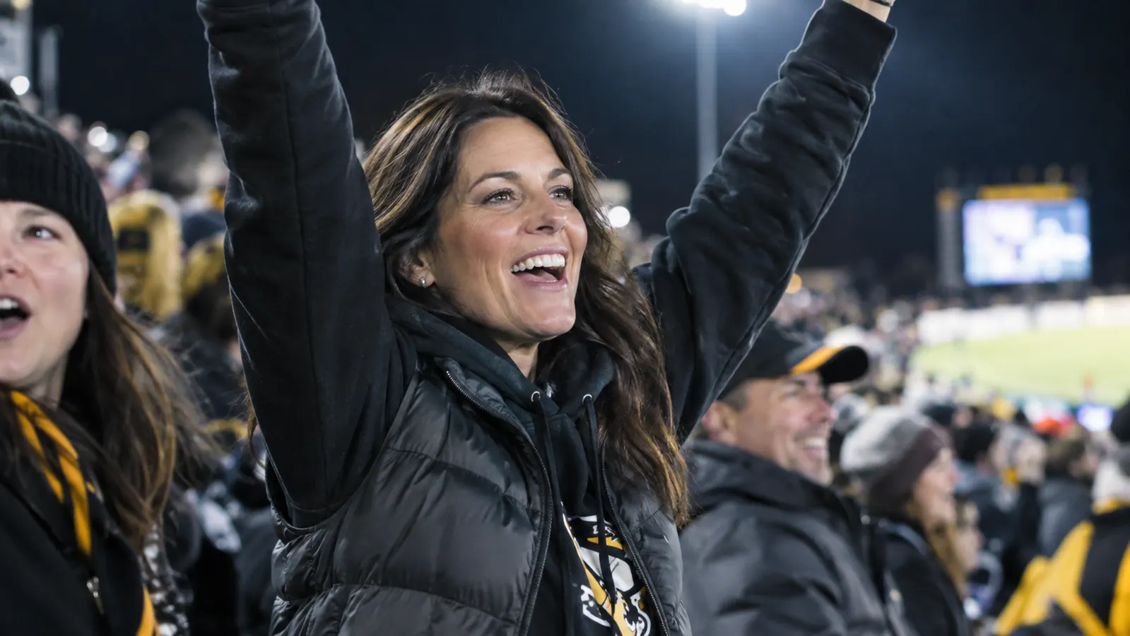 A woman in a team hoodie and a lightweight vest cheering in the stands at a night football game