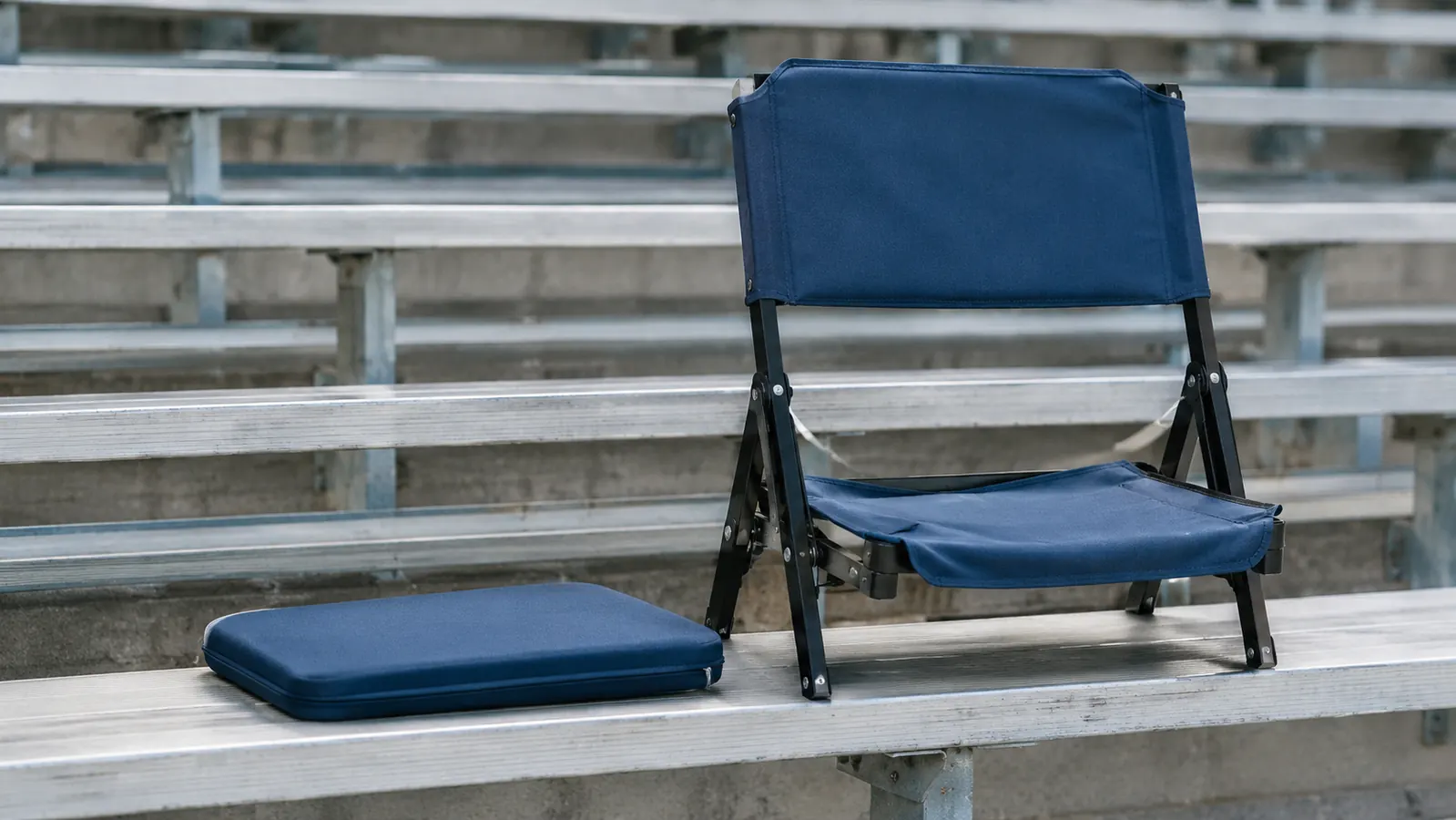 A flat foam cushion on an empty bleacher bench beside a folding stadium seat with a backrest