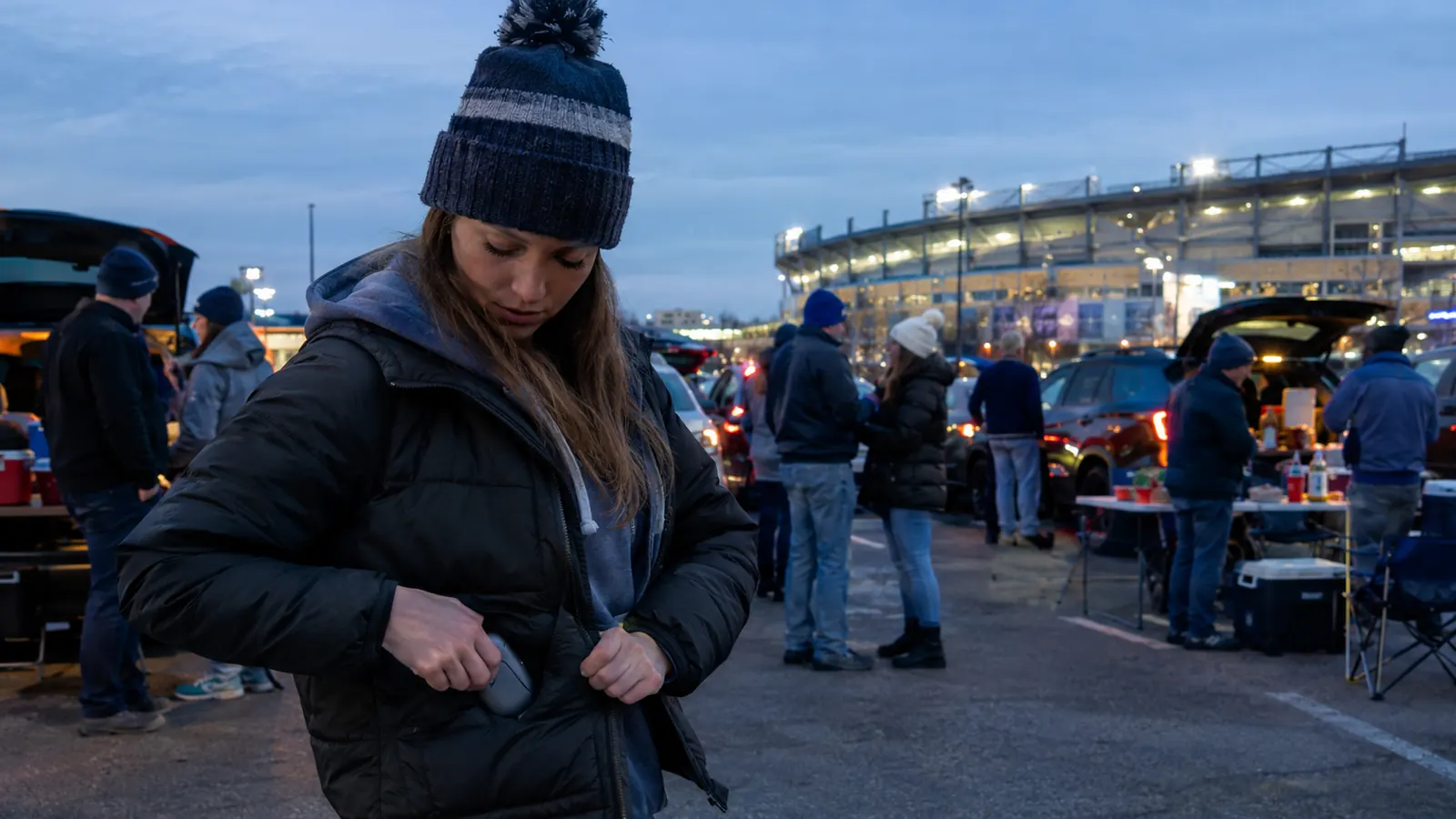 Fans tailgating at dusk in a stadium parking lot, one woman slipping a small warmer into her jacket pocket
